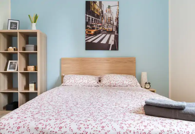 Chambre lumineuse et bien aménagée avec grand lit double, couette à motif, table de chevet avec lampe et réveil, et étagère décorative — idéale pour les annonces mettant en avant le confort et la lumière naturelle.