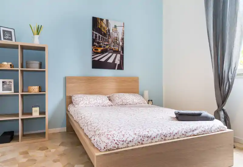 Chambre lumineuse et soignée avec un lit en bois, une literie florale et une étagère cubique ; la lumière naturelle et le cadre mural créent une atmosphère chaleureuse.