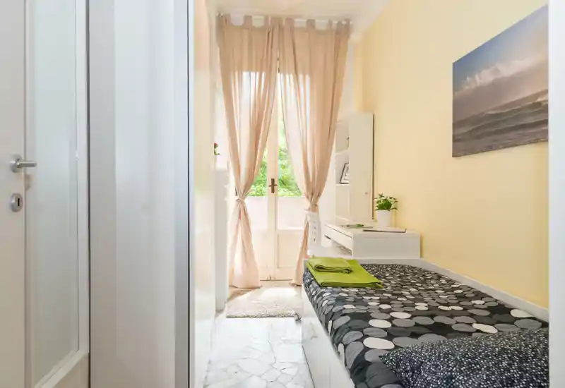 Chambre simple compacte avec lit bien fait, bureau et étagères à côté d’une porte-fenêtre donnant sur un petit balcon. La lumière naturelle tamisée par des rideaux légers et des tons neutres avec des accents verts créent une ambiance calme et accueillante.
