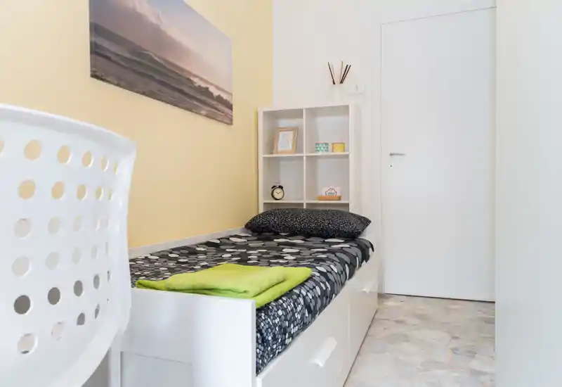 Chambre simple compacte avec lit bien fait, étagères décoratives et lumière naturelle douce — lumineuse, propre et accueillante.