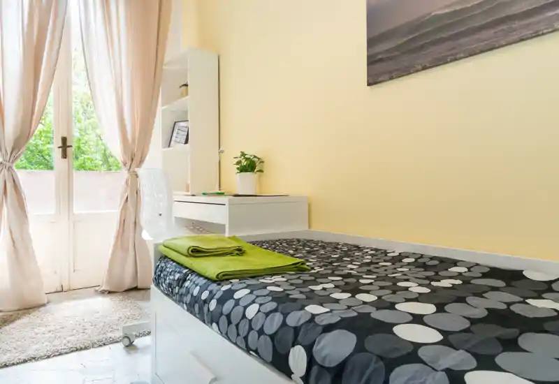 Chambre compacte et lumineuse avec un lit simple au linge à motifs, des serviettes pliées et un petit bureau près de la porte du balcon laissant entrer la lumière naturelle.