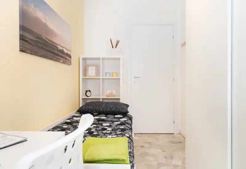 Neat single bedroom with a patterned bed, bright walls and minimalist shelving. The room is well lit and styled with a simple desk area, wall art and folded linens on the bed.