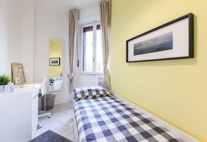 Dormitorio individual compacto y luminoso con edredón de cuadros, escritorio junto a la ventana y detalles decorativos neutros que generan una sensación fresca y ordenada.