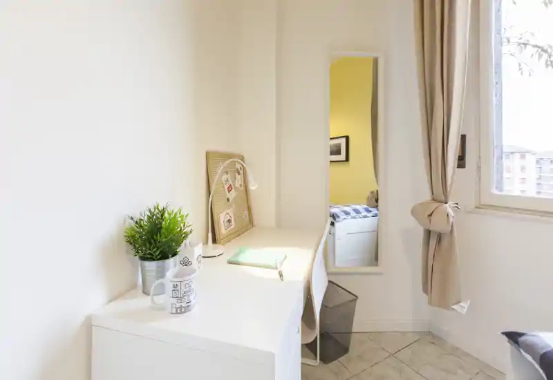 Rincón de dormitorio compacto y luminoso con escritorio blanco y un espejo estrecho que refleja parte de la cama; estilo moderno y luz natural desde la ventana.