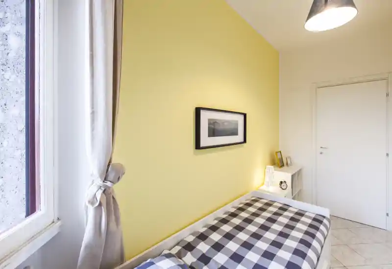 Dormitorio pequeño y luminoso con cama individual de ropa a cuadros, pared de acento amarilla, cuadro enmarcado y mesita con lámpara — ambiente ordenado y acogedor para alquileres de corta estancia.