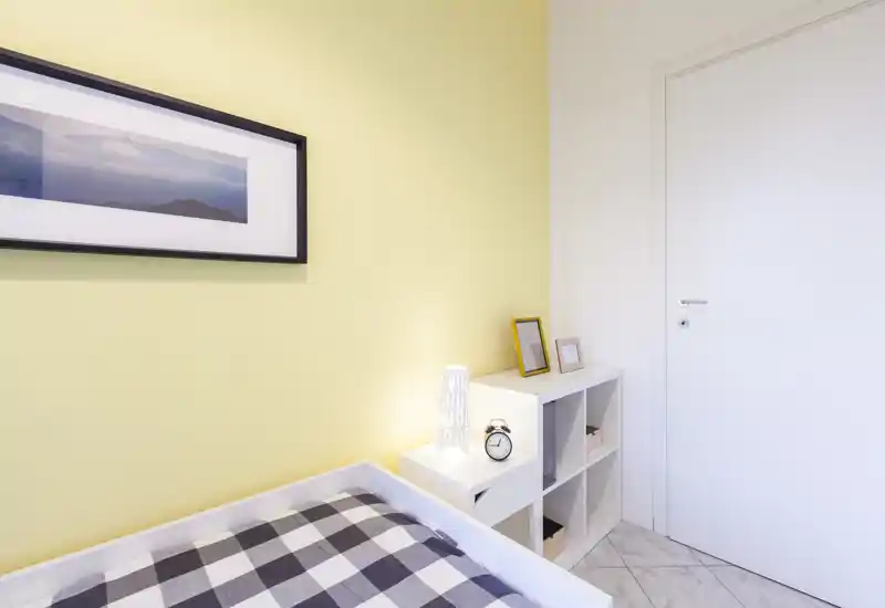 Dormitorio individual luminoso y minimalista con colcha a cuadros, lámpara de mesita y pequeño estante de cubos — paredes en tonos pastel y cuadro sencillo que crean un ambiente ordenado y sereno.