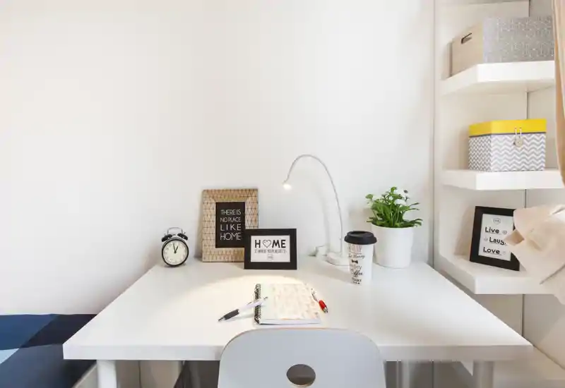Spazio di lavoro luminoso e minimal con scrivania bianca, lampada da lettura, cornici decorative e una piccola pianta — perfetto per studio o smart working.