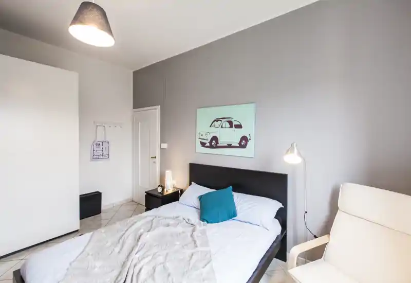 Camera da letto moderna e luminosa con letto matrimoniale, comodino e lampada da lettura. Lenzuola pulite, un cuscino decorativo turchese e una stampa murale creano un’atmosfera tranquilla e accogliente adatta agli annunci.