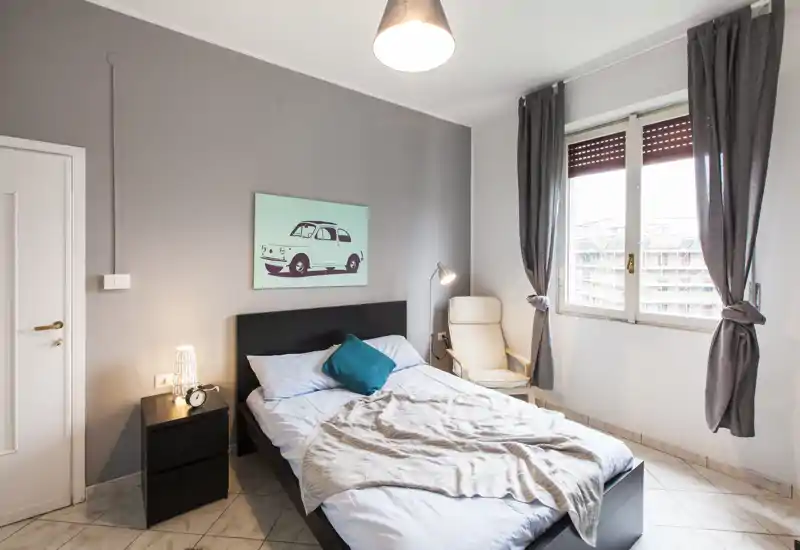 Camera da letto luminosa e moderna con letto matrimoniale, comodino e una poltrona vicino alla finestra; tessuti morbidi e toni neutri creano un ambiente accogliente.