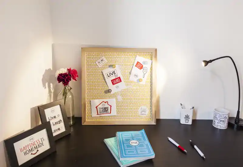 Zona de escritorio/vanity ordenada con cuadros, tablero de corcho, cuadernos y lámpara de mesa — luminosa y bien decorada.