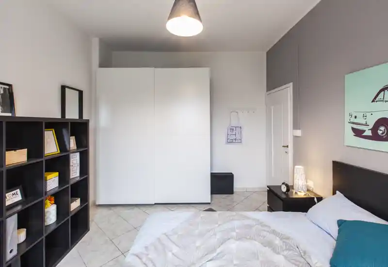 Camera da letto moderna e luminosa con grande armadio bianco, libreria a cubi e letto ben rifatto con cuscino accentato; lampada e quadro sulla parete creano un’atmosfera tranquilla e accogliente.