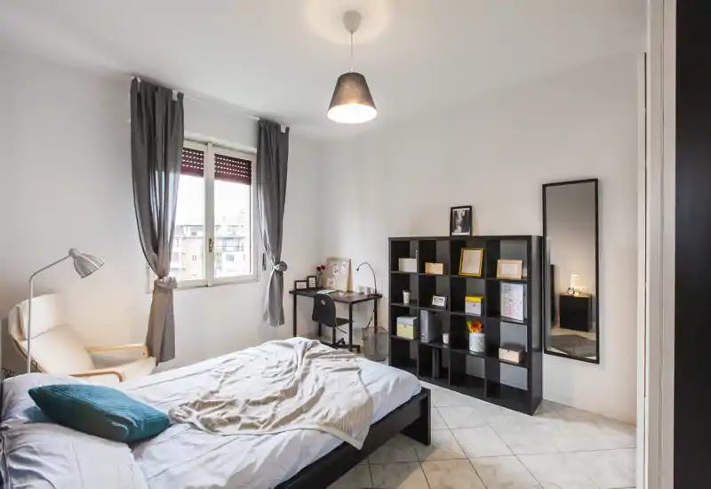 Camera da letto luminosa e ben arredata con letto matrimoniale, poltrona e una scrivania vicino alla finestra; libreria e specchio a figura intera completano lo spazio.