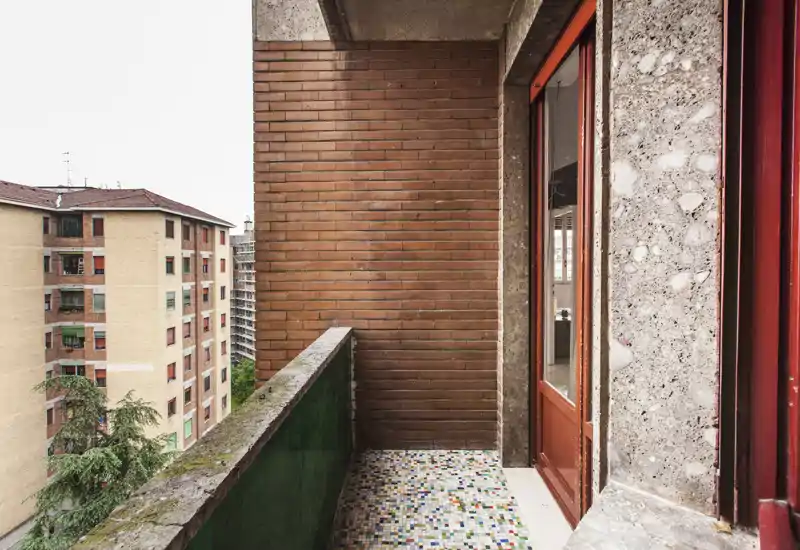 Pequeño balcón urbano con suelo de baldosas decoradas, pared de ladrillo y barandilla verde; vista a edificios residenciales cercanos. Ideal para un rincón exterior pequeño.