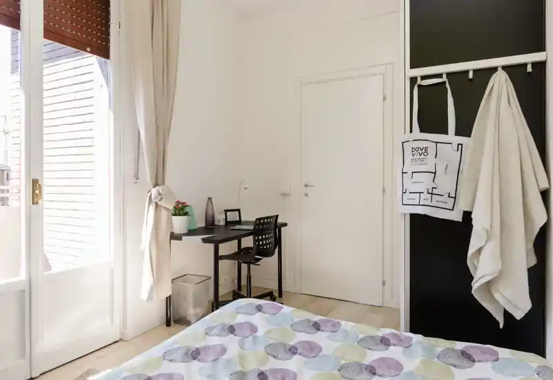 Dormitorio luminoso y bien decorado con edredón estampado, escritorio junto a la puerta del balcón y agradable luz natural—ideal para alquileres temporales.