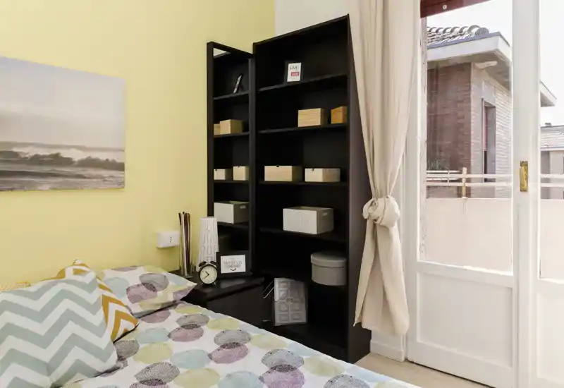 Rincón de dormitorio luminoso y acogedor con ropa de cama estampada, cojines decorativos y una estantería alta junto a una gran puerta de cristal que aporta luz natural.