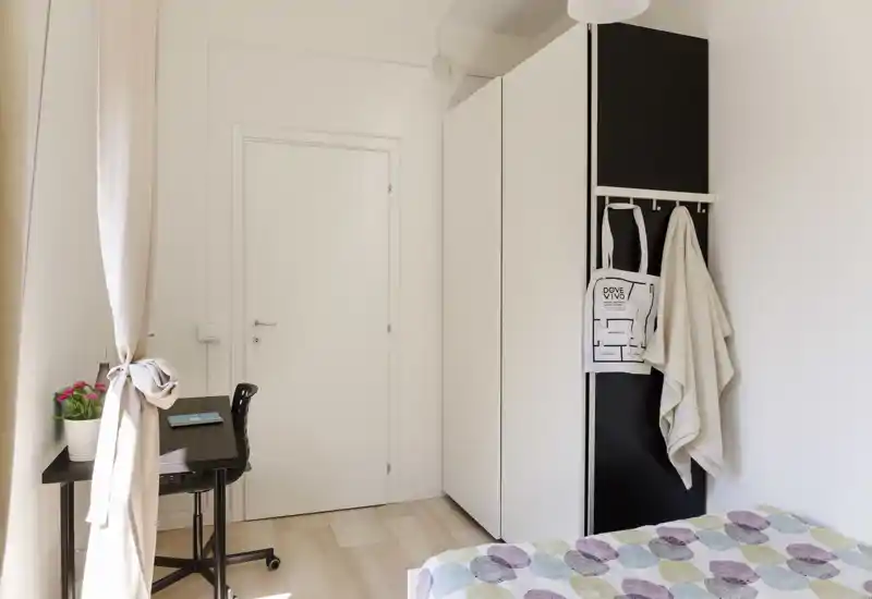 Dormitorio compacto y luminoso con colcha estampada, armario alto blanco con perchas y un pequeño escritorio junto a la ventana — estilo minimalista cuidado y mucha luz natural.