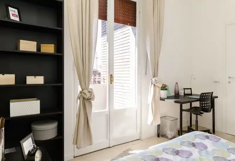 Rincón de dormitorio luminoso y bien decorado con colcha estampada, escritorio y gran puerta acristalada que aporta luz natural.