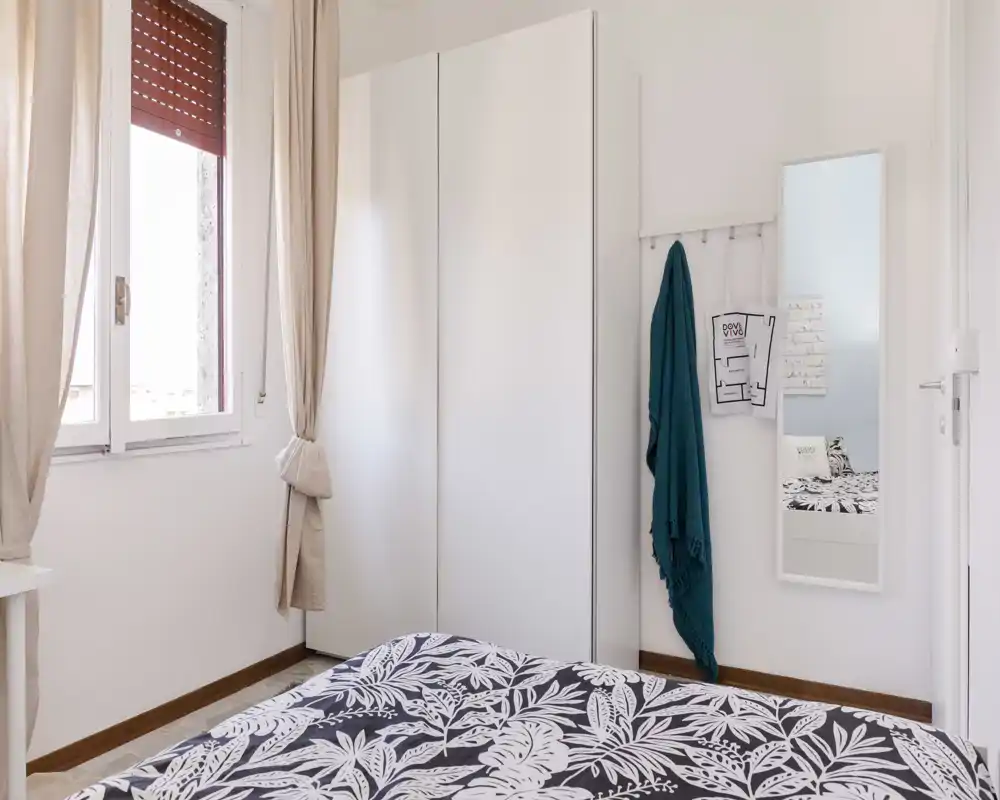 Dormitorio luminoso y minimalista con edredón estampado, gran armario blanco y luz natural desde la ventana. La composición destaca la zona de la cama y la decoración limpia, ideal para un anuncio de alquiler.
