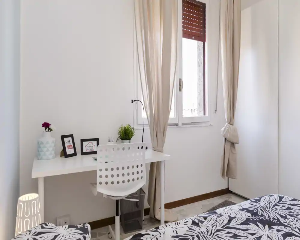 Dormitorio luminoso y de estilo sencillo con colcha estampada, escritorio junto a la ventana y cortinas neutras que crean un ambiente acogedor.