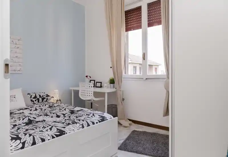 Dormitorio luminoso y bien presentado con cama de marco blanco, ropa de cama estampada y un pequeño escritorio junto a la ventana — ideal para una persona o como habitación de invitados.