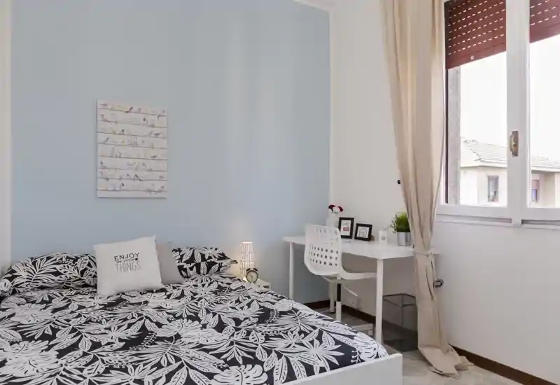 Dormitorio luminoso y ordenado con cama doble, ropa de cama estampada, lámpara de mesilla y pequeño escritorio junto a la ventana, ideal para descansar y trabajar ligeramente.