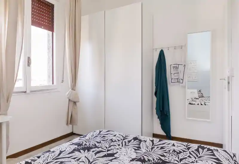 Dormitorio luminoso y minimalista con edredón estampado, gran armario blanco y luz natural desde la ventana. La composición destaca la zona de la cama y la decoración limpia, ideal para un anuncio de alquiler.