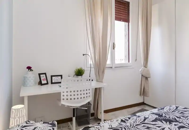 Dormitorio luminoso y de estilo sencillo con colcha estampada, escritorio junto a la ventana y cortinas neutras que crean un ambiente acogedor.