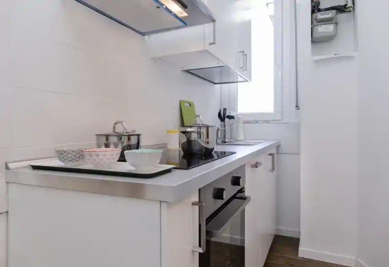 Cocina compacta moderna y luminosa con vitrocerámica, horno empotrado y utensilios en la encimera; muebles blancos y luz natural por la ventana.