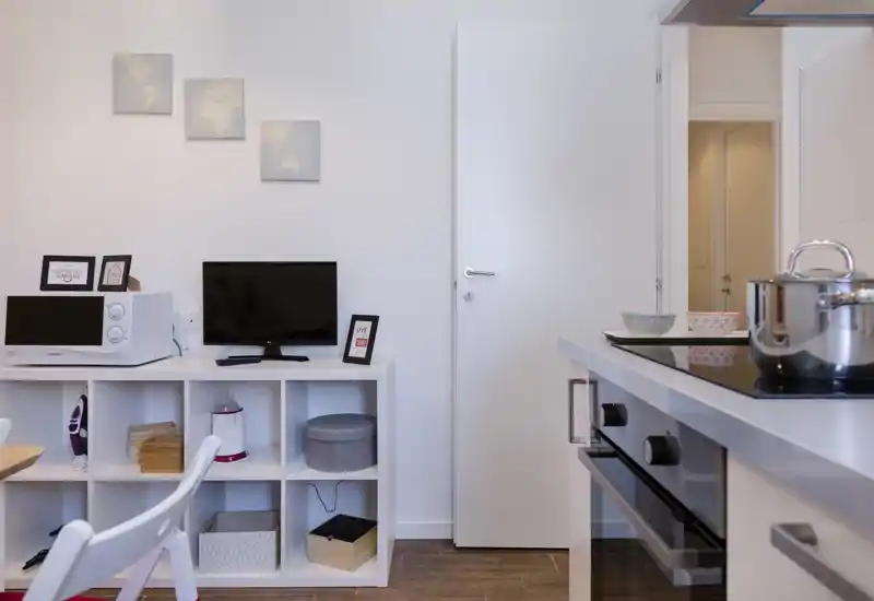 Cocina americana compacta con placa de inducción y horno en primer plano, y una estantería con microondas y televisión. Estilo moderno y limpio, paredes claras y suelo de madera, ideal para un apartamento pequeño.