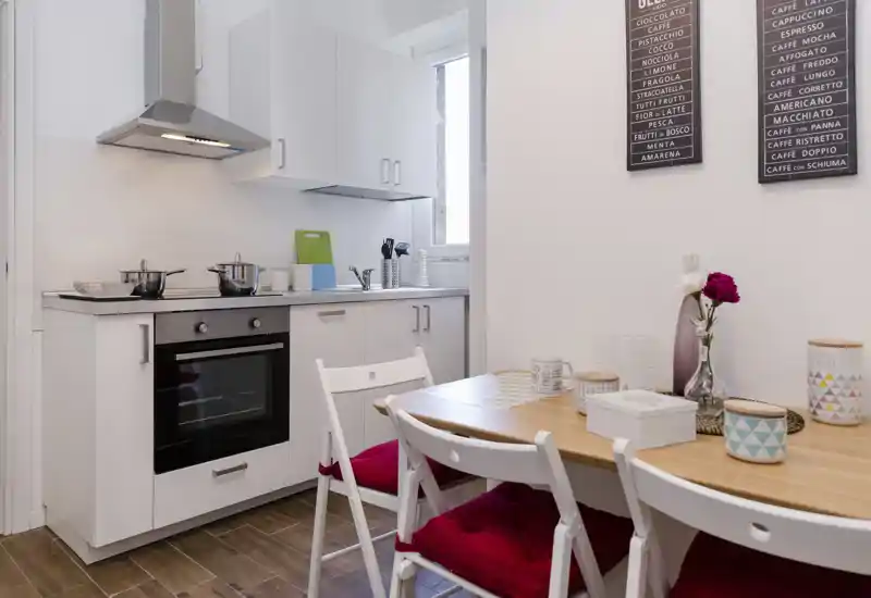Cocina compacta y moderna con muebles blancos, horno empotrado, campana de acero y una mesa de madera con cojines rojos — luminosa y bien preparada para alquileres vacacionales.
