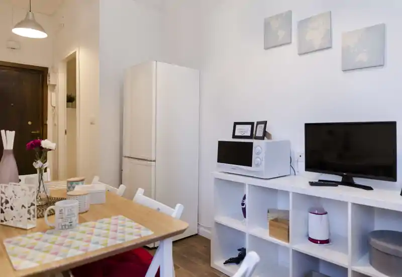 Cocina-comedor compacta y luminosa con mesa de madera preparada para comer, frigorífico y electrodomésticos pequeños en un ambiente moderno y acogedor.