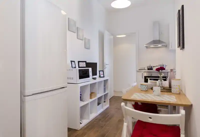 Cocina compacta y moderna con suelo efecto madera, pequeña mesa de comedor con cojines rojos y vajilla decorativa, horno empotrado y campana — luminosa y acogedora para un piso urbano.