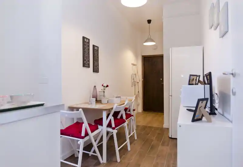 Pequeña zona de comedor con mesa estrecha de madera, cuatro sillas plegables blancas con cojines rojos y suelos de madera cálida. Espacio limpio y bien iluminado con detalles decorativos sencillos, ideal como rincón de comedor en un apartamento.