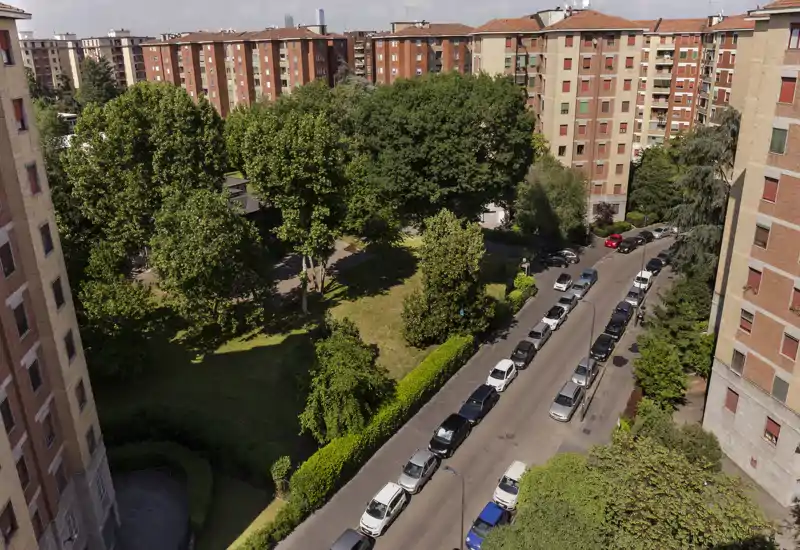 Vista exterior luminosa de un patio residencial con árboles frondosos, césped verde y coches aparcados en una calle tranquila — ideal para mostrar los espacios verdes y el entorno del edificio.