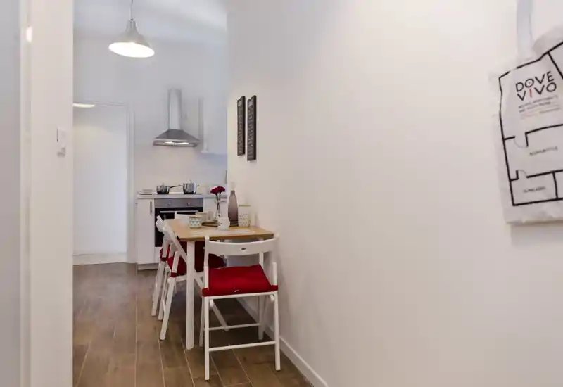 Kitchenette compacta y luminosa con una pequeña mesa de madera y dos sillas blancas con cojines rojos; campana y cocina modernas al fondo.