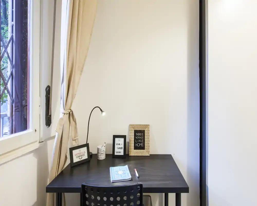 Bright, tidy workspace with a black desk by a window, small lamp and framed decorative prints — a cozy study corner ideal for focused work.