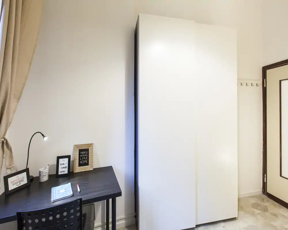 Compact room with a black writing desk, chair, decorative frames and a tall white wardrobe. Neutral walls and soft curtains make the space bright and functional — ideal to describe a small bedroom or study corner.