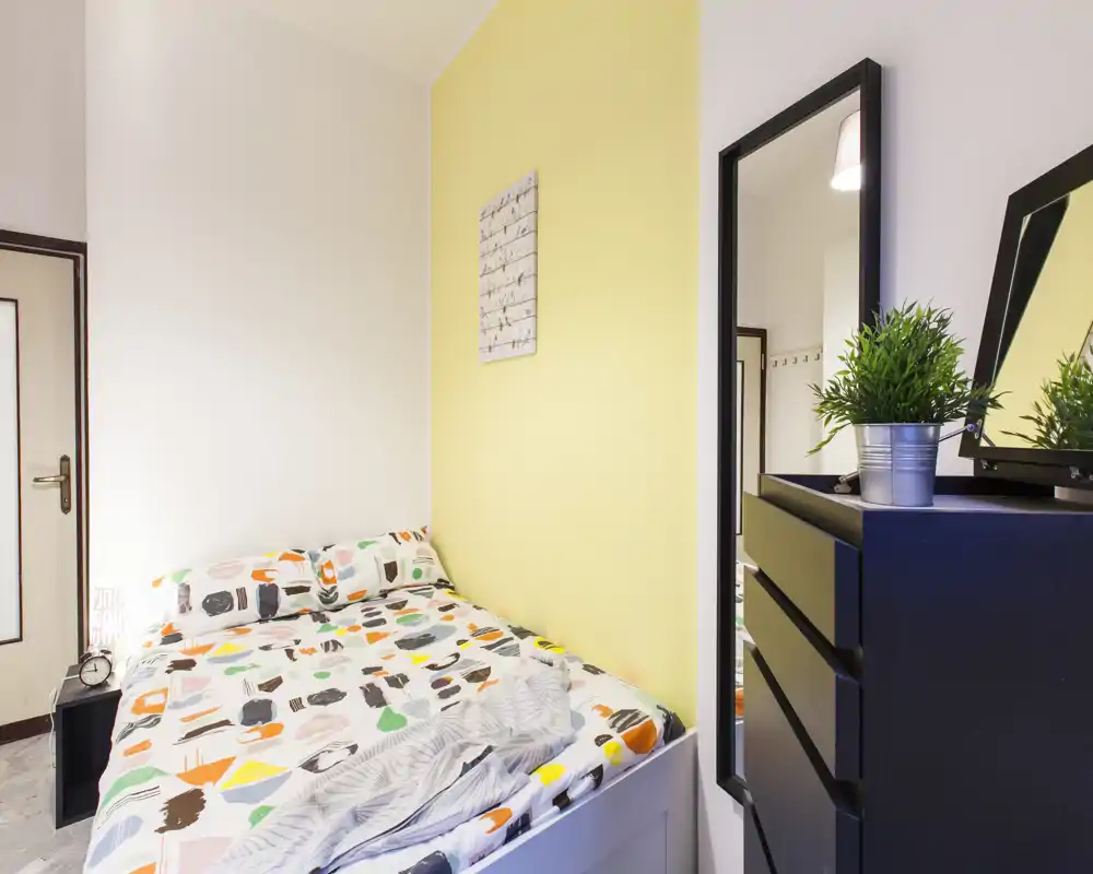 Bright, compact bedroom with a patterned double bed pushed against a soft yellow accent wall, a tall mirror and a chest of drawers with a small plant.