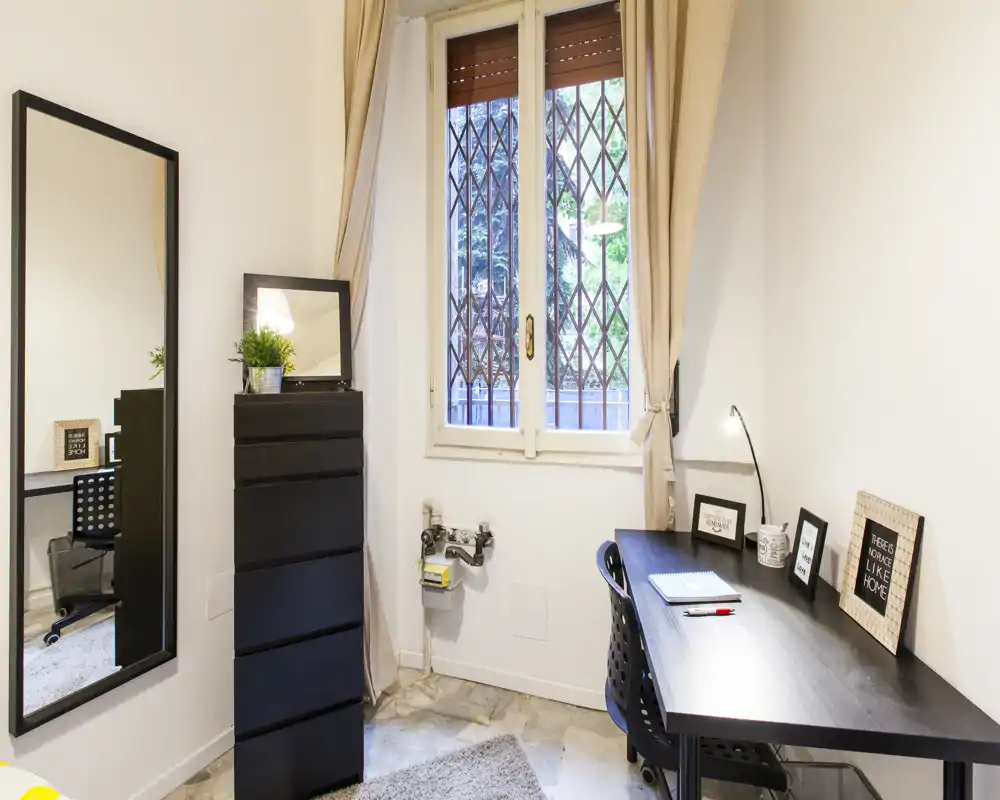 Bright, compact home office corner with a desk, chair, lamp and decorative frames beside a window; neutral tones and a tall mirror make the small room feel larger.