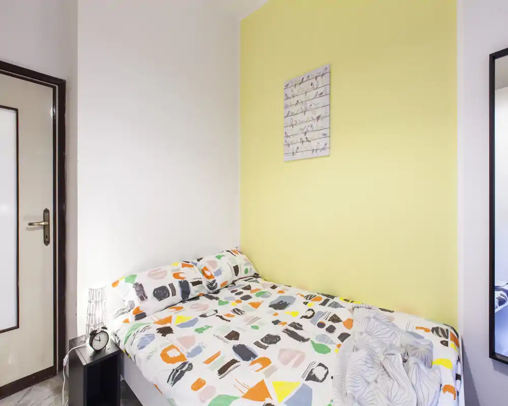 Bright, compact bedroom with patterned bedding, bedside lamp and minimalist wall art — ideal for a single or couple. The room is neatly staged with cheerful yellow accent wall and clear, even lighting.