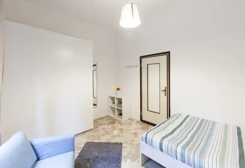 Bright, compact bedroom with a single bed dressed in striped linens, a small sofa, wardrobe and a rug — clean minimalist styling ideal for a solo tenant.