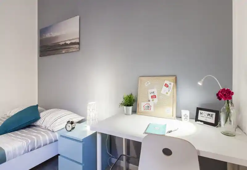 Bright, modern bedroom corner with a neatly made single bed and a tidy white desk. Soft grey walls, decorative accents and a desk lamp create a cozy workspace next to the bed — ideal for a student or single occupant.