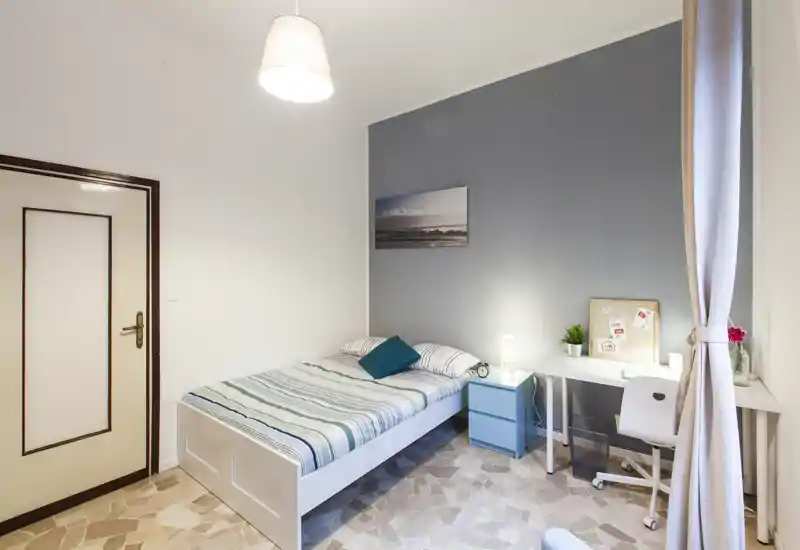 Camera singola luminosa e ordinata con letto a righe, comodino e una scrivania vicino alla finestra — ideale per uno studente o un affittuario singolo.