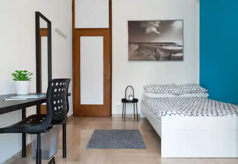 Dormitorio individual moderno y luminoso con ropa de cama estampada, mesita y pequeño escritorio. Estilo minimalista y pared azul de acento que crean un espacio fresco y acogedor, perfecto para alquileres.