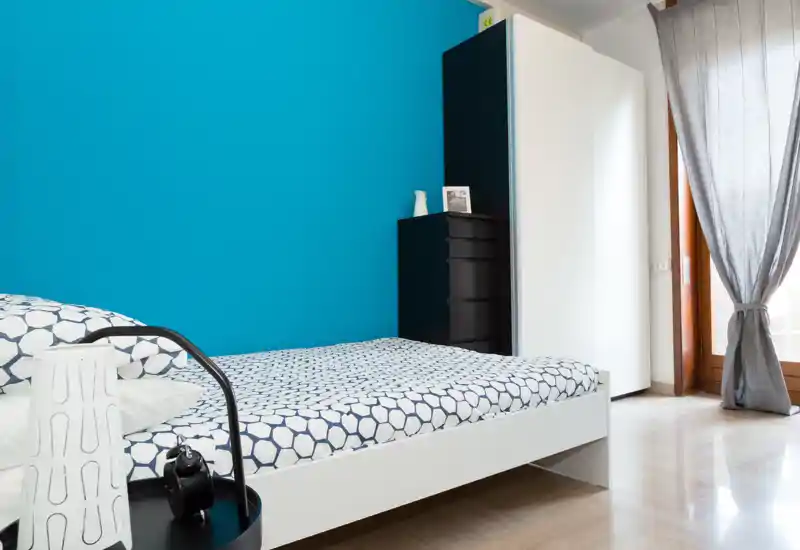 Dormitorio moderno y luminoso con ropa de cama estampada y una pared azul turquesa; gran armario y puerta a balcón con cortina que aporta luz natural.