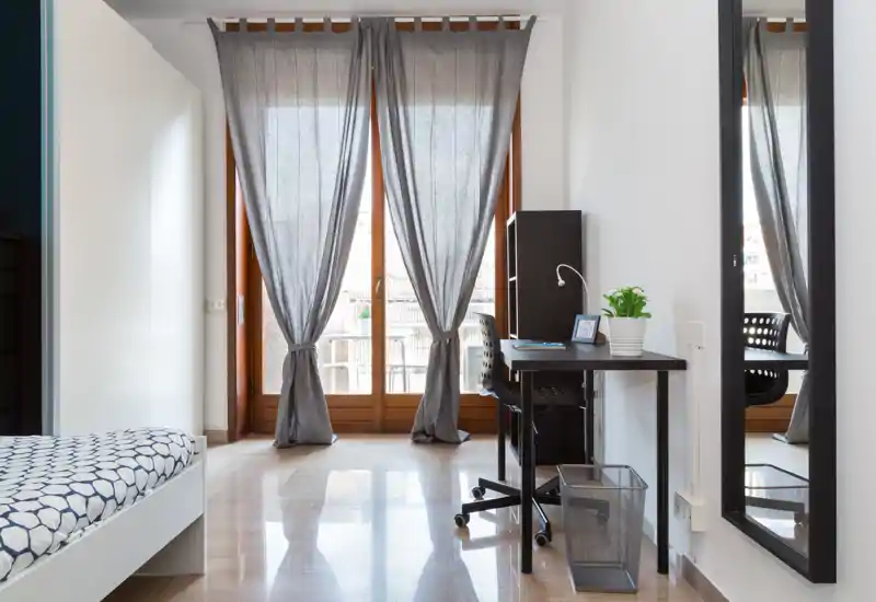 Dormitorio moderno y luminoso con cama individual a la izquierda, escritorio y silla junto a las puertas del balcón y cortinas grises que dejan pasar mucha luz natural.