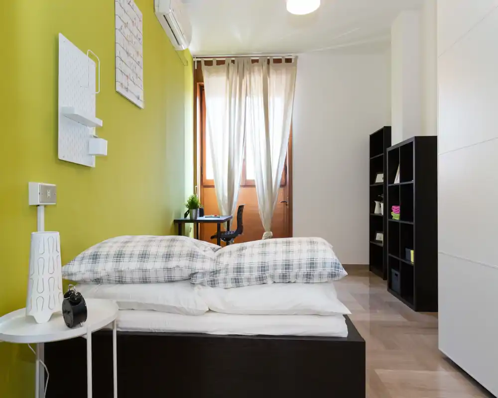 Camera da letto moderna e luminosa con un letto matrimoniale ben rifatto in primo piano, parete accentuata verde, scrivania vicino alla finestra e scaffalatura nera.