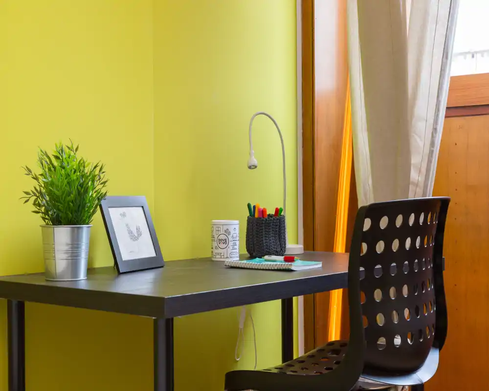 Angolo studio luminoso e compatto con scrivania nera moderna e sedia forata, cancelleria ordinata, una piccola lampada, stampa incorniciata e una pianta in vaso su una parete verde chiaro — ideale per lavoro o studio.