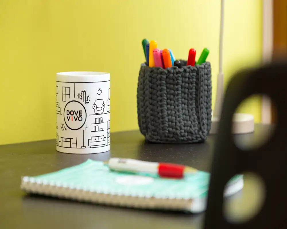 Primo piano di un piano d'appoggio in zona giorno con un portapenne, un piccolo contenitore brandizzato e un blocco note; parete gialla calda e luce soffusa creano un'atmosfera accogliente.