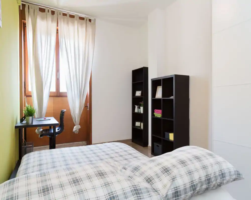 Camera da letto luminosa e ordinata con un grande letto in primo piano, biancheria a motivi, scrivania vicino alla finestra e scaffalature aperte per riporre.